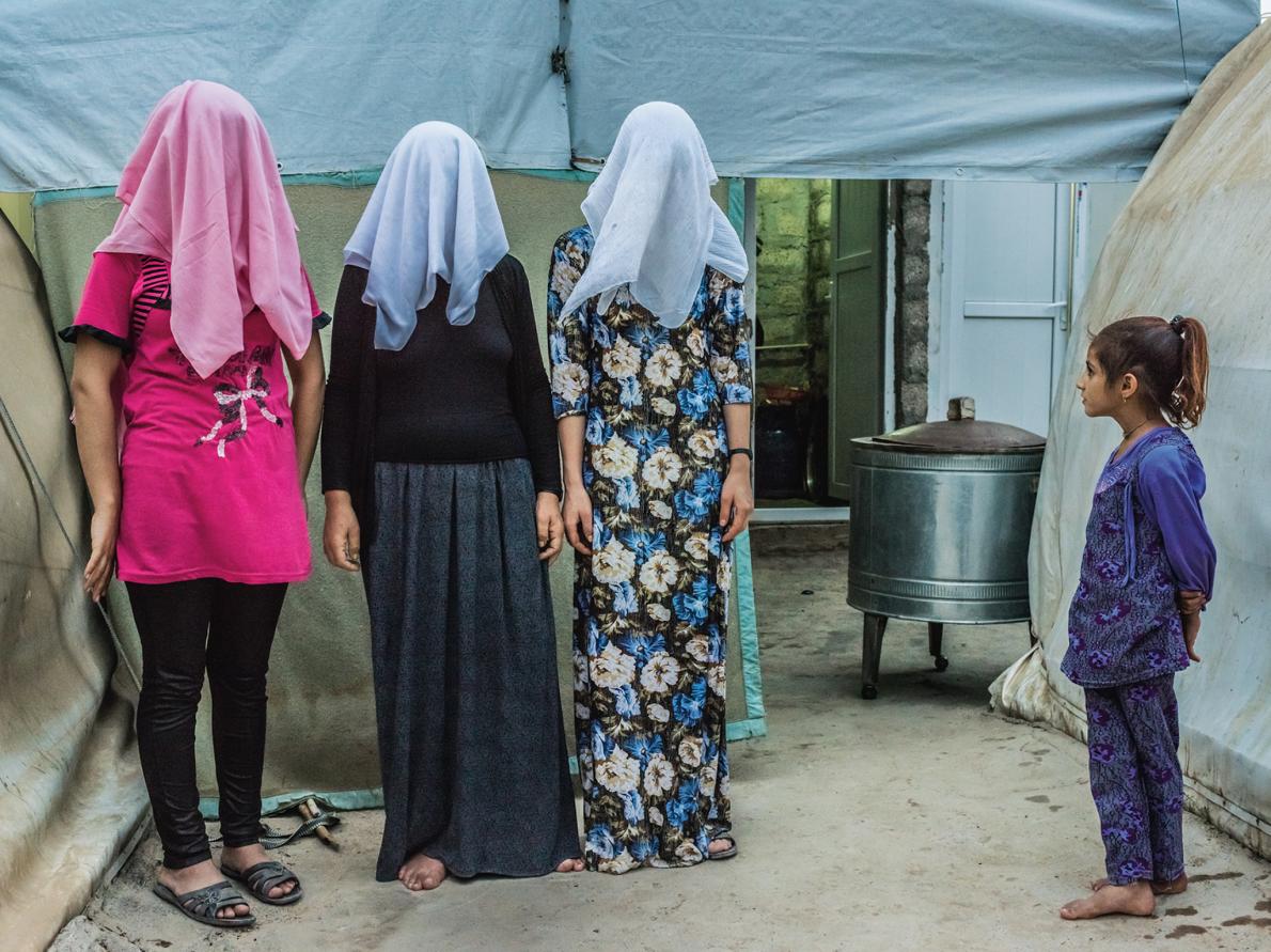 Một cô gái đứng nhìn trong khi ba người phụ nữ người Kurd đang chụp ảnh với khuôn mặt của họ ẩn. Hai phụ nữ nói rằng họ đã buộc phải kết hôn với máy bay chiến đấu ISIS trước khi chạy trốn đến một trại tị nạn.